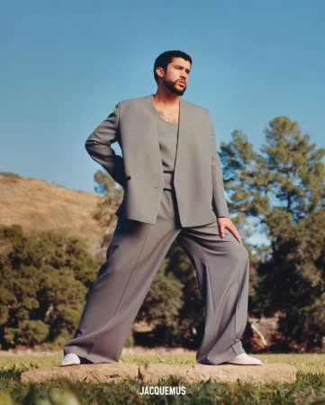 Bad Bunny wearing a grey suit and matching top standing outdoors on low stone blocks against a clear blue sky and grassy hills.