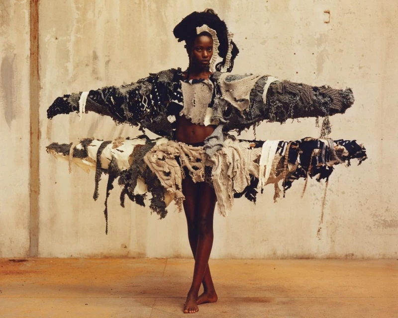 Striking studio shot of a dark-skinned fashion model in a large, textural couture garment with a neutral background, posing barefoot.