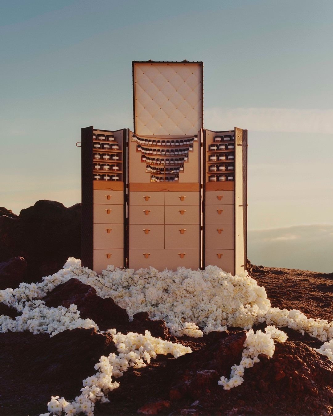 Tall wooden dresser with drawers and compartments on a hill, juxtaposed against a light blue sky, creating a surreal outdoor scene.