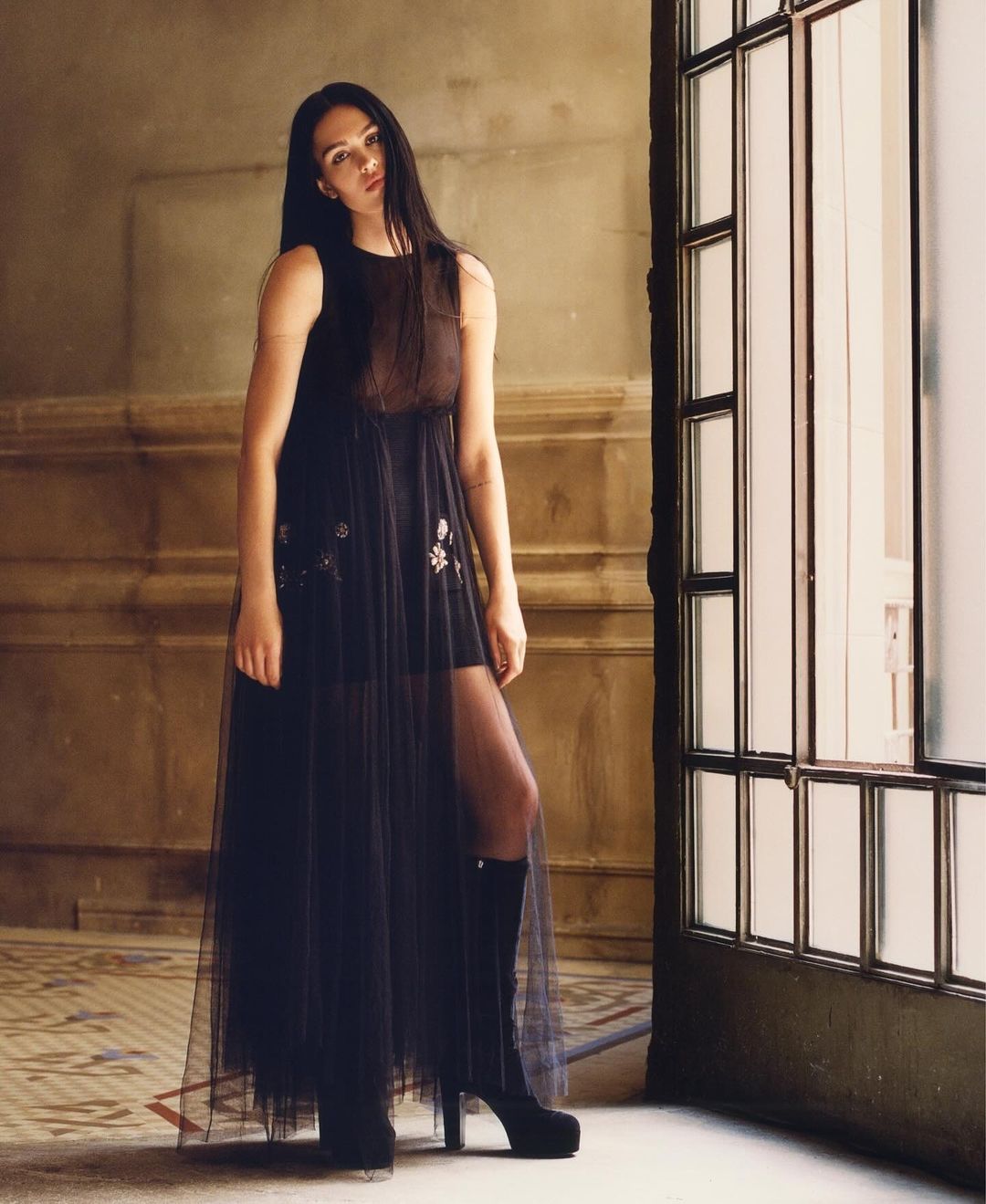 Fashionable woman in a sheer black dress and black boots poses in an elegant interior by a window, with tiled floors and neutral walls.