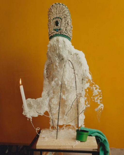 Surreal still life photography featuring a mannequin-like sculpture with ornate crown, candle, green pot, and towel against an orange background.