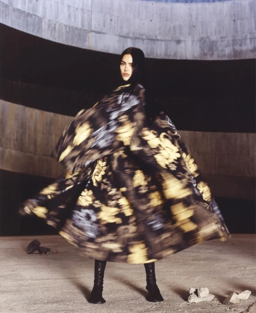 A woman in a floral patterned dress poses in an indoor concrete setting, with a blurred effect suggesting movement, and wearing boots.