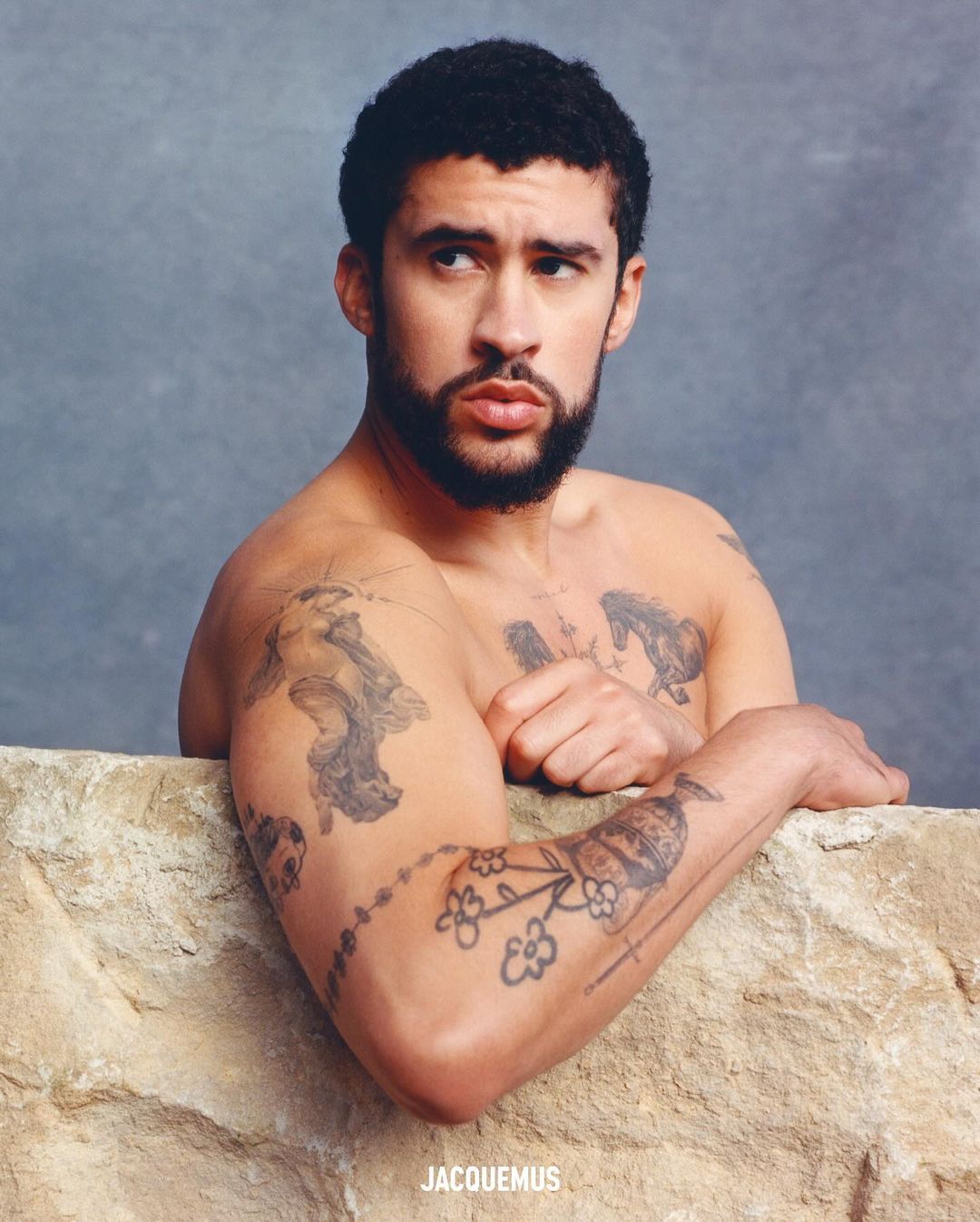 Portrait of shirtless adult male with tattoos, posed in front of a textured stone wall and grey background with soft studio lighting.
