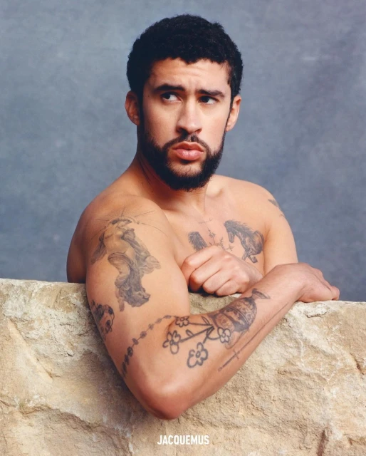 Portrait of shirtless adult male with tattoos, posed in front of a textured stone wall and grey background with soft studio lighting.