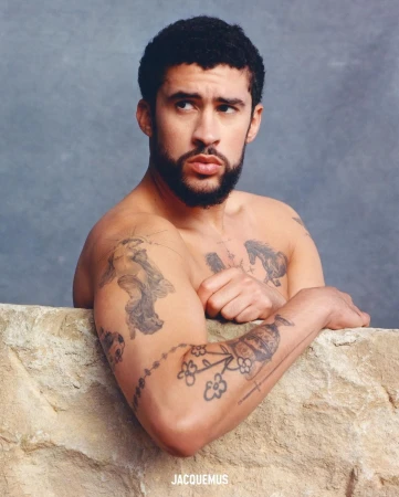 Portrait of shirtless adult male with tattoos, posed in front of a textured stone wall and grey background with soft studio lighting.