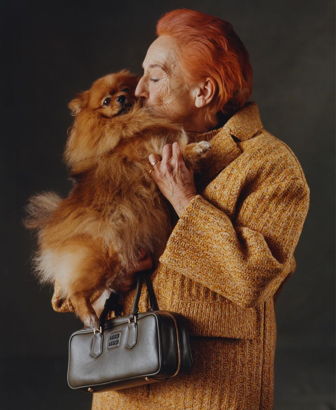 Elderly woman with orange hair in a yellow coat holds a fluffy orange Pomeranian dog and a dark handbag in a studio setting.