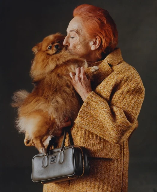 Elderly woman with orange hair in a yellow coat holds a fluffy orange Pomeranian dog and a dark handbag in a studio setting.