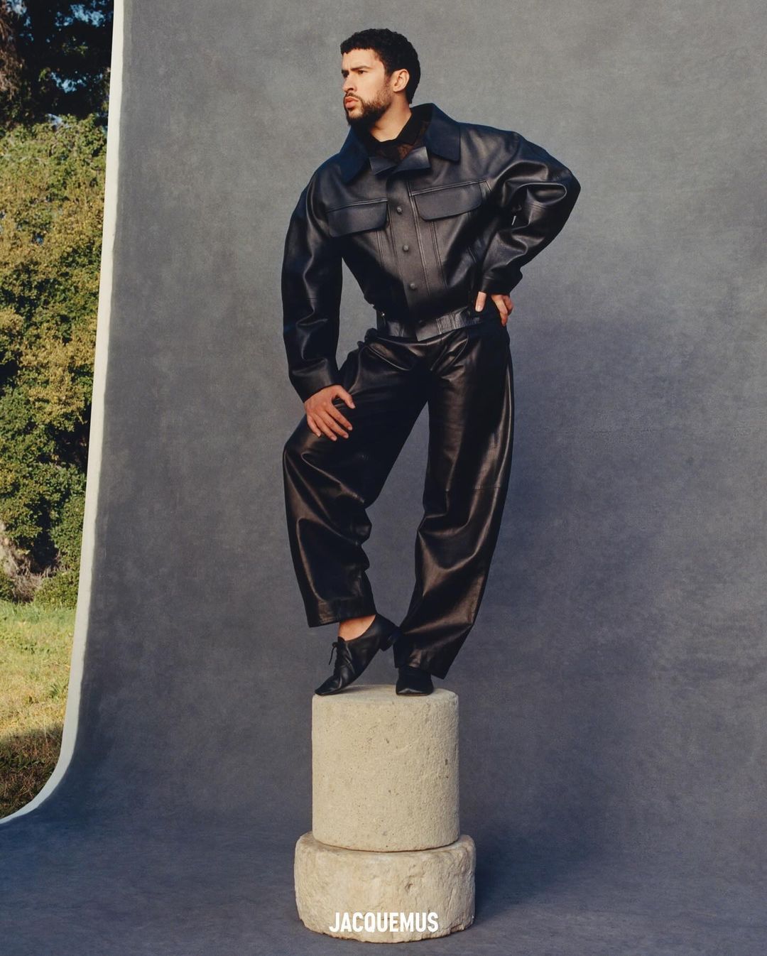 A man in a black leather jacket and pants poses on stacked concrete cylinders against a neutral backdrop in a fashion editorial.