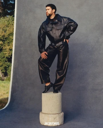 A man in a black leather jacket and pants poses on stacked concrete cylinders against a neutral backdrop in a fashion editorial.