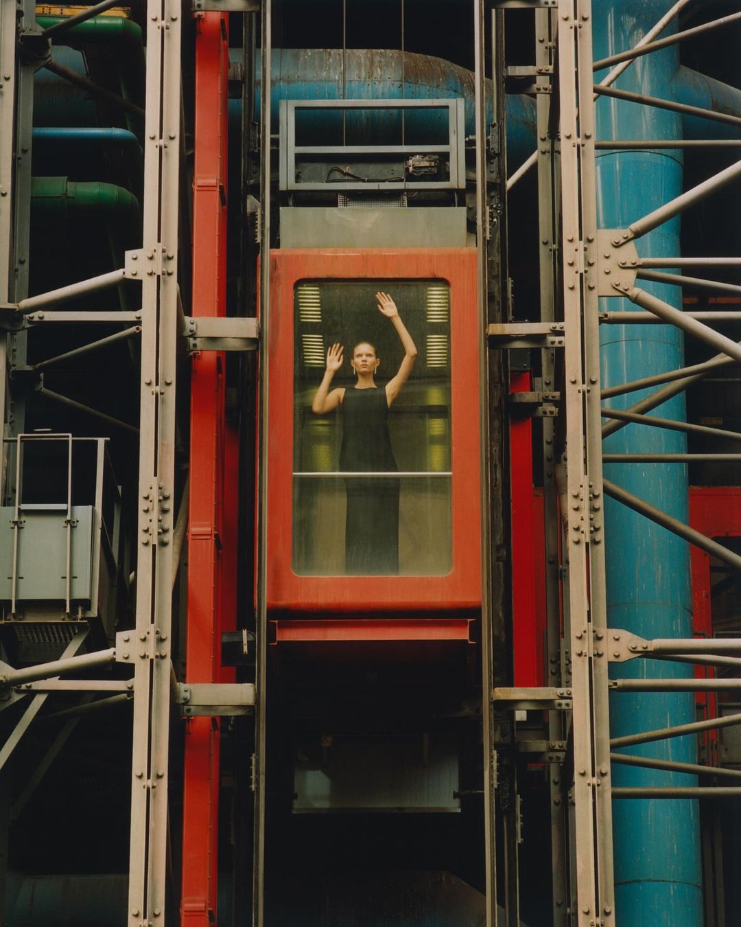 A woman in a black dress is inside a red metal and glass elevator, holding up her hands, with architectural metal beams and pipes around it.