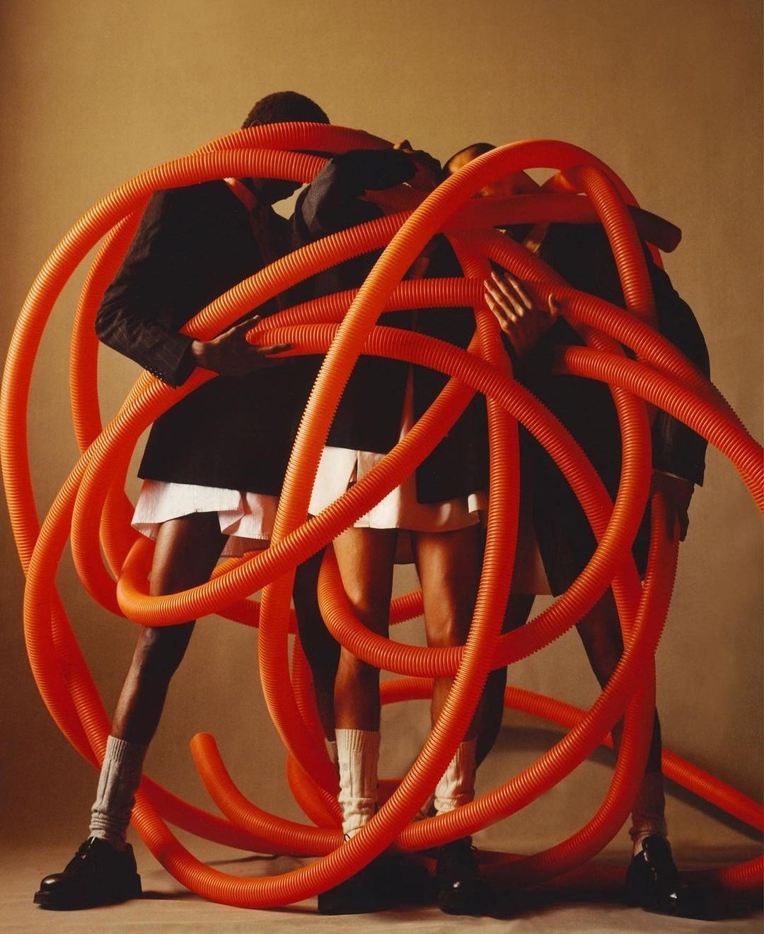 Three models wrapped in large, orange corrugated tubes wearing dark blazers, white bottoms, and black shoes posing in a studio setting.