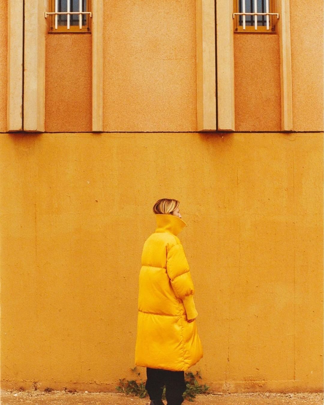 A woman in a yellow coat stands against a matching yellow wall with architectural details. The image features a monochrome color scheme.