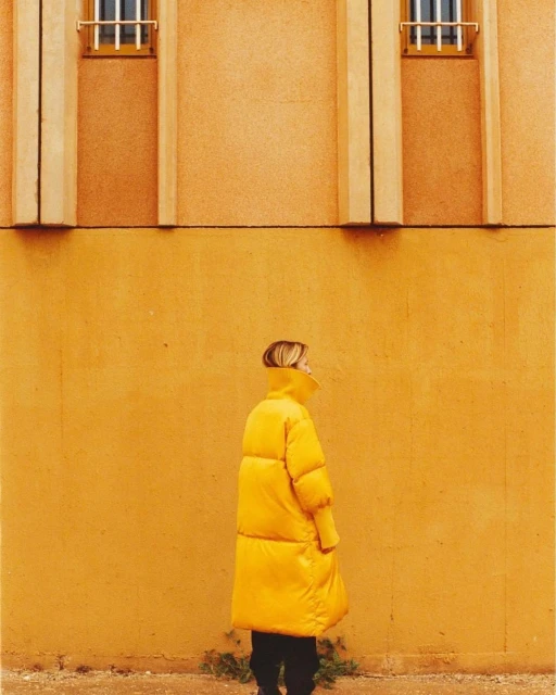 A woman in a yellow coat stands against a matching yellow wall with architectural details. The image features a monochrome color scheme.