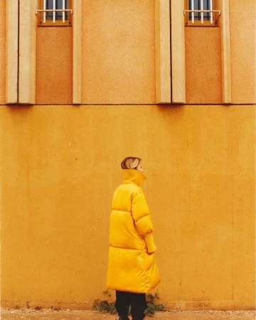 A woman in a yellow coat stands against a matching yellow wall with architectural details. The image features a monochrome color scheme.