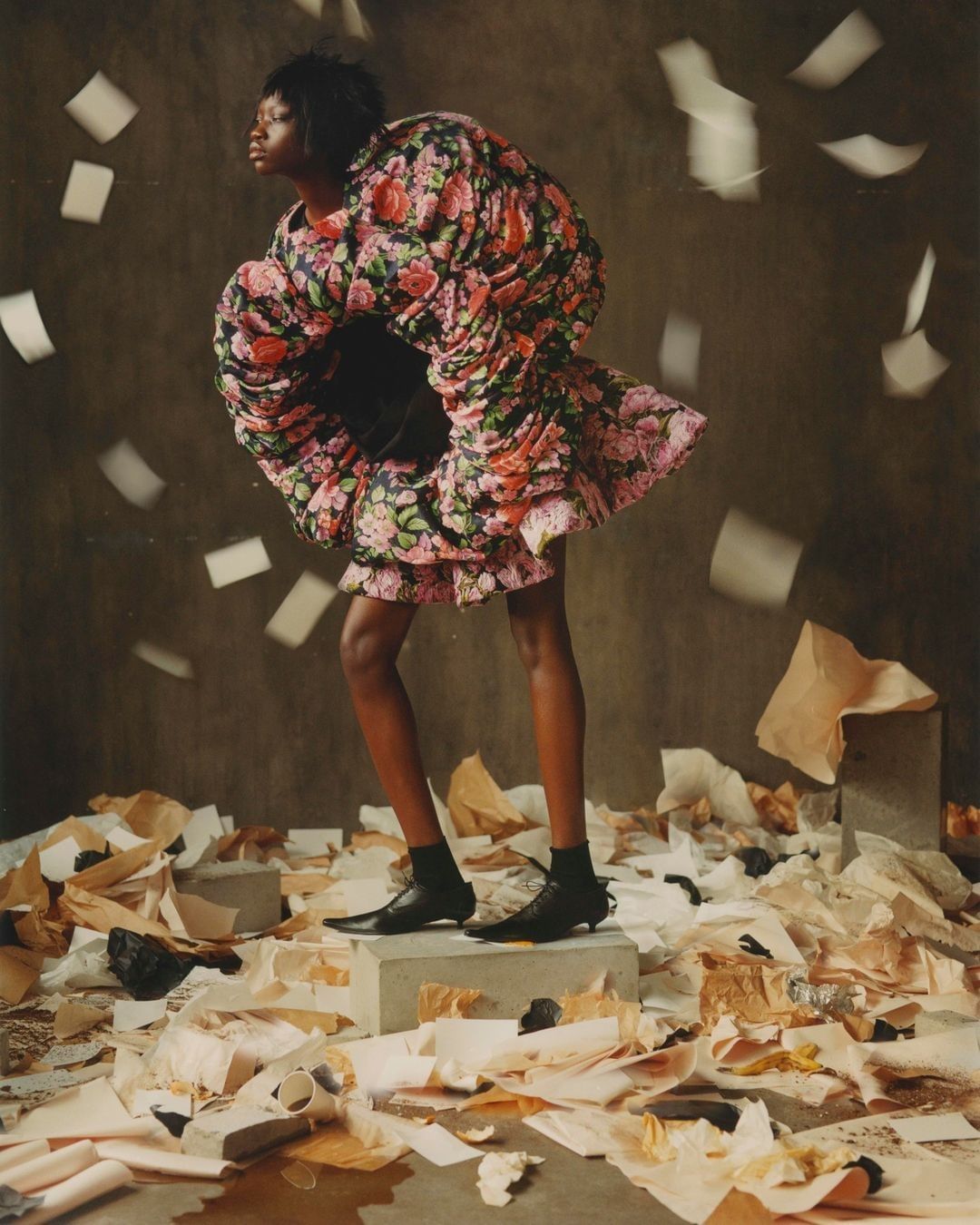 Woman in floral dress stands on concrete block amidst scattered papers and falling pages in a fashion editorial studio shot.