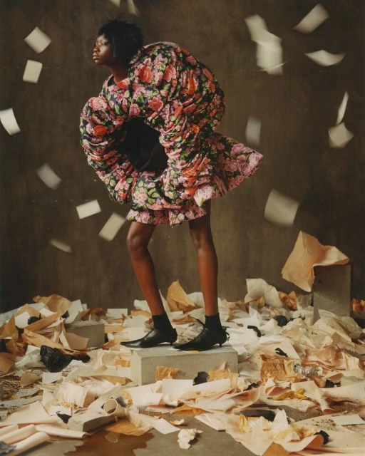 Woman in floral dress stands on concrete block amidst scattered papers and falling pages in a fashion editorial studio shot.