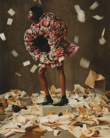 Woman in floral dress stands on concrete block amidst scattered papers and falling pages in a fashion editorial studio shot.