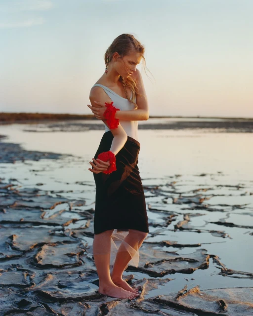 Woman in a light-colored bodysuit and black skirt poses on a cracked, dry lakeshore at sunset with red flowers on her arms.