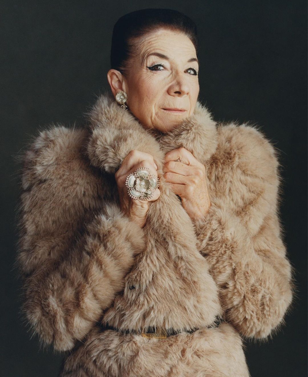 Older woman in a fur coat and large ring strikes a pose in a studio setting, creating a classic portrait with neutral background.