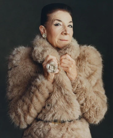 Older woman in a fur coat and large ring strikes a pose in a studio setting, creating a classic portrait with neutral background.