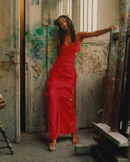 A stylish adult female poses in a red dress inside an artist studio, with paint-splattered walls and floors adding to the creative ambiance.