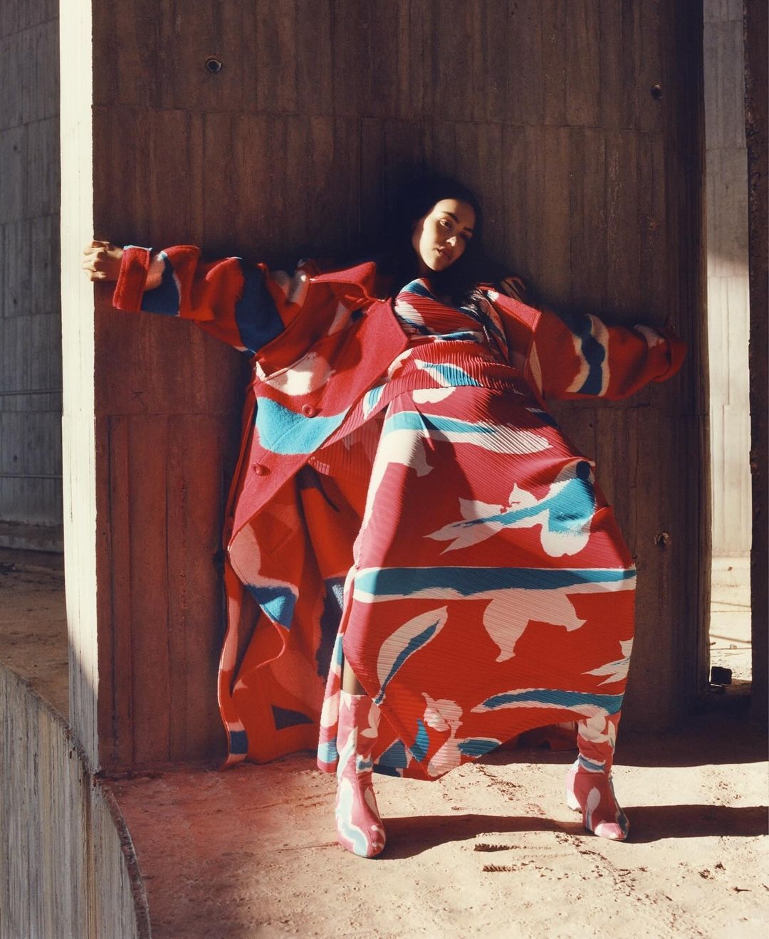 A woman poses in a red patterned coat and matching boots, leaning against a concrete wall in an urban setting. Fashion photography.
