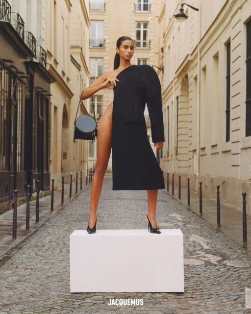 Fashion shot of a stylish woman in a black coat and heels, posing on a white box in a narrow urban street with tall buildings.
