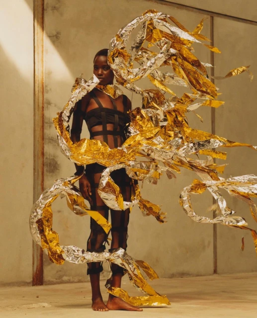 Fashion shot of a Black woman in a black leotard with gold and silver ribbon-like accents, standing indoors against a plain wall.