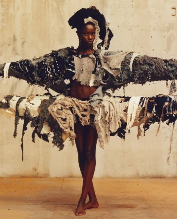 A Black woman posing in an avant-garde textured sculptural garment against a weathered concrete wall background.