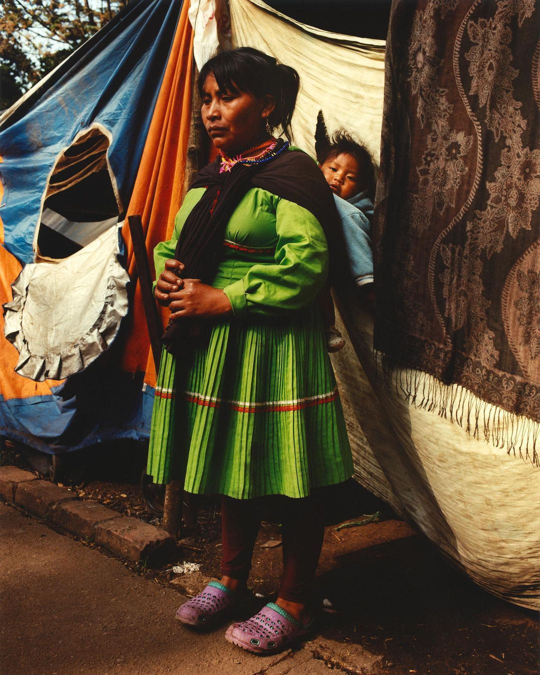 Indigenous woman and child in traditional clothing stand in front of a patterned tent, creating a vibrant cultural portrait.