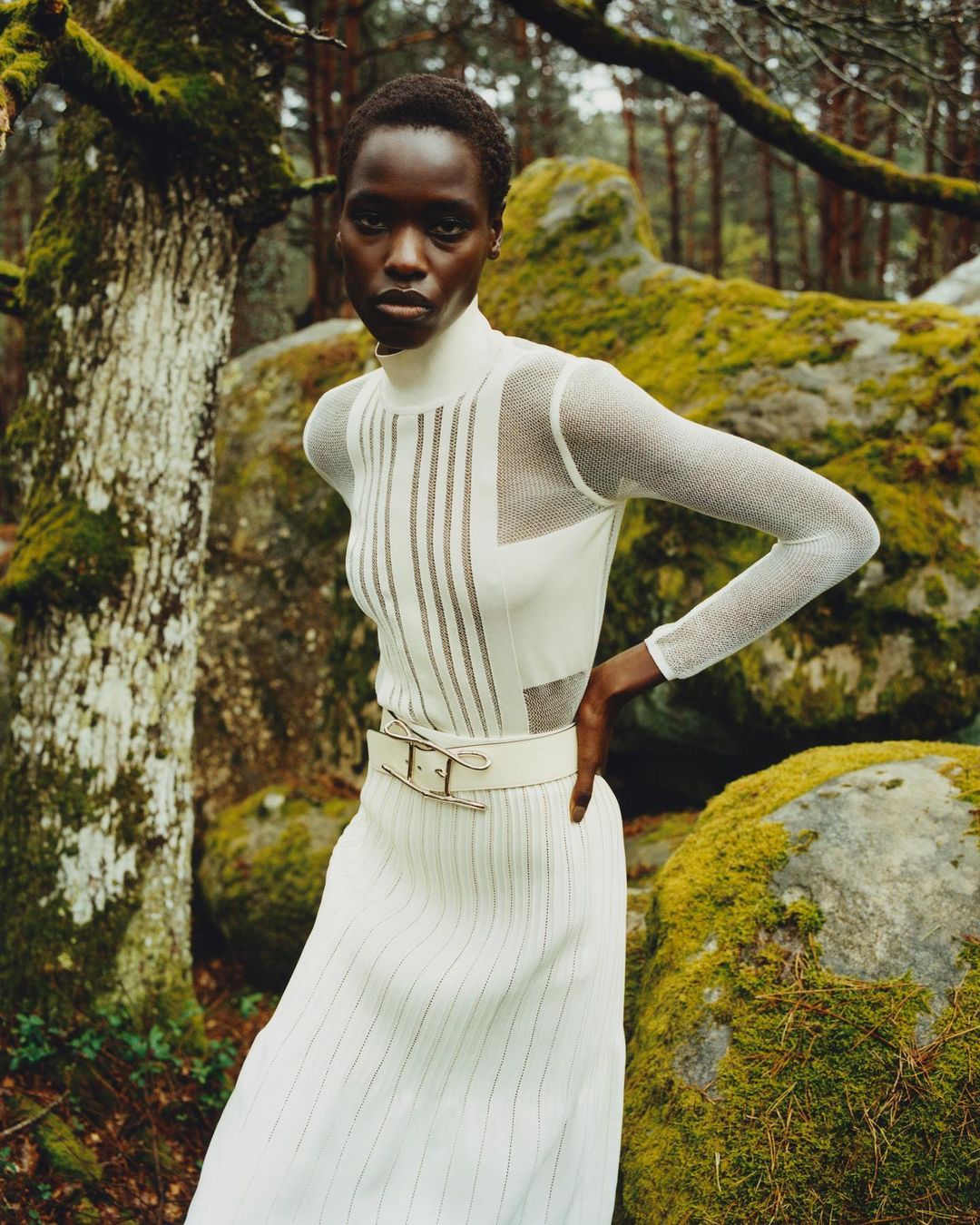 Fashion shot of an adult Black female model in a white dress with mesh sleeves posing in a forest with moss-covered rocks in the background.