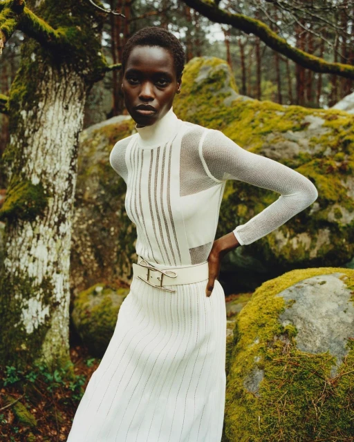 Fashion shot of an adult Black female model in a white dress with mesh sleeves posing in a forest with moss-covered rocks in the background.