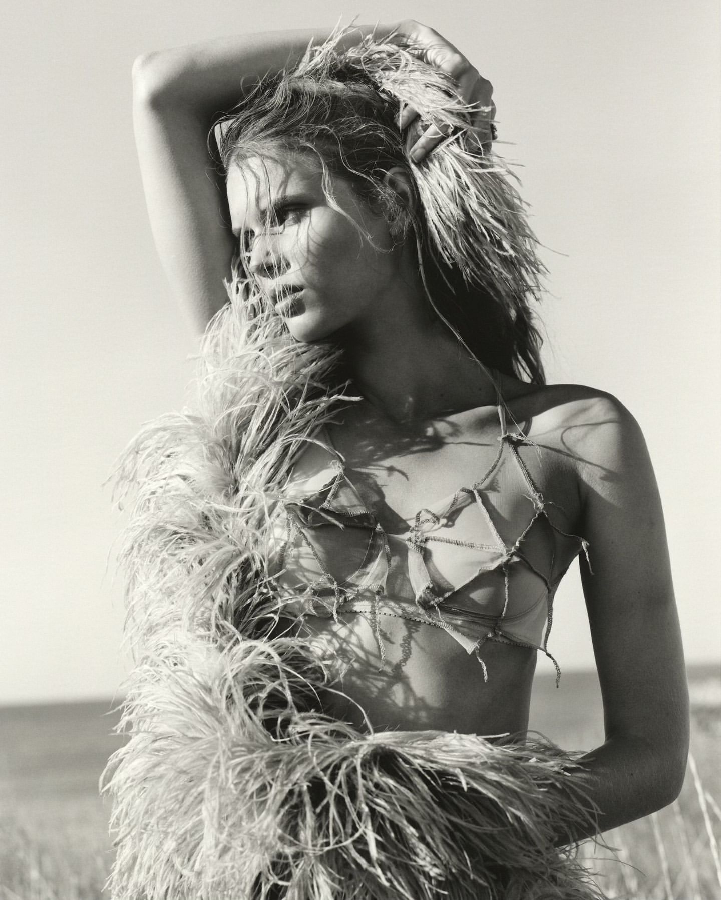 Black and white portrait of a woman outdoors with a feathery garment, eyes glancing left, posing in natural light against a neutral sky.