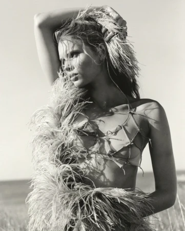 Black and white portrait of a woman outdoors with a feathery garment, eyes glancing left, posing in natural light against a neutral sky.
