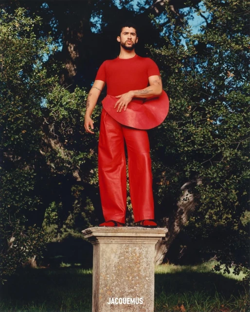 Man in red outfit stands on stone pedestal in outdoor fashion shot featuring Jacquemus branding, set against trees and blue sky.