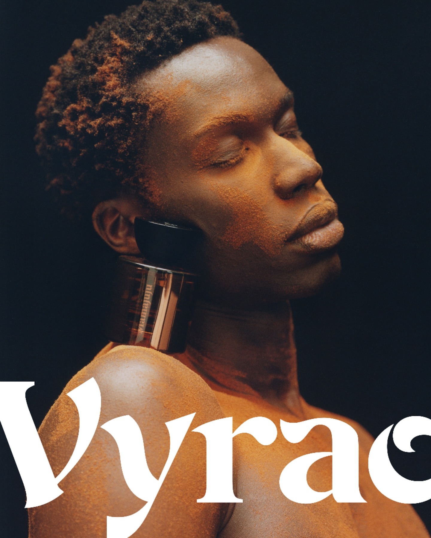 Close-up studio shot of a shirtless Black male with bronze body paint, eyes closed, and a perfume bottle resting on his shoulder against a dark backdrop.