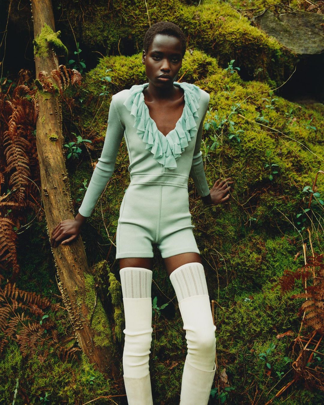 Adult woman in light green shorts and top with ruffled neckline, stands near a moss-covered fallen tree in a verdant forest setting.