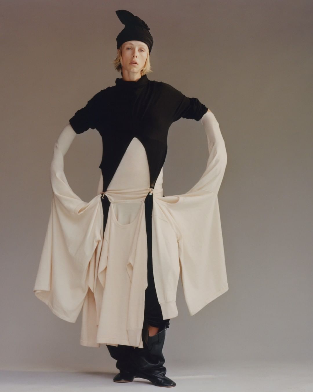 Fashionable woman in a black top, unique light-beige skirt with suspended shirt layers, black hat, and boots posing in a studio.