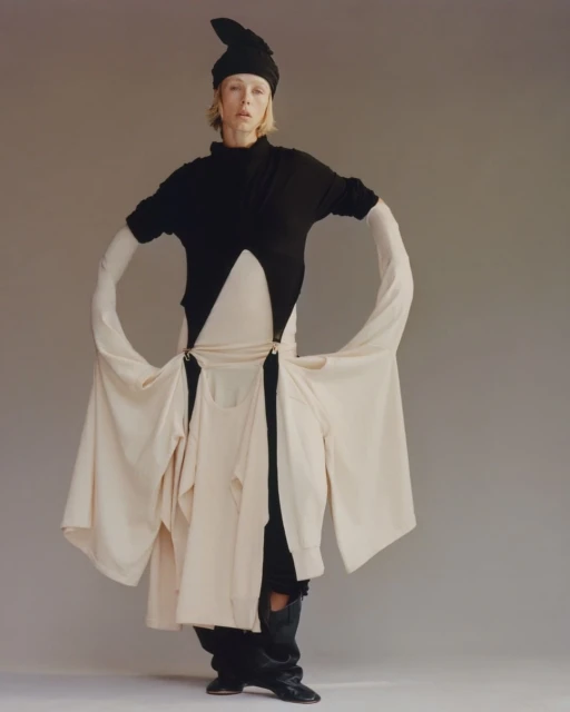 Fashionable woman in a black top, unique light-beige skirt with suspended shirt layers, black hat, and boots posing in a studio.