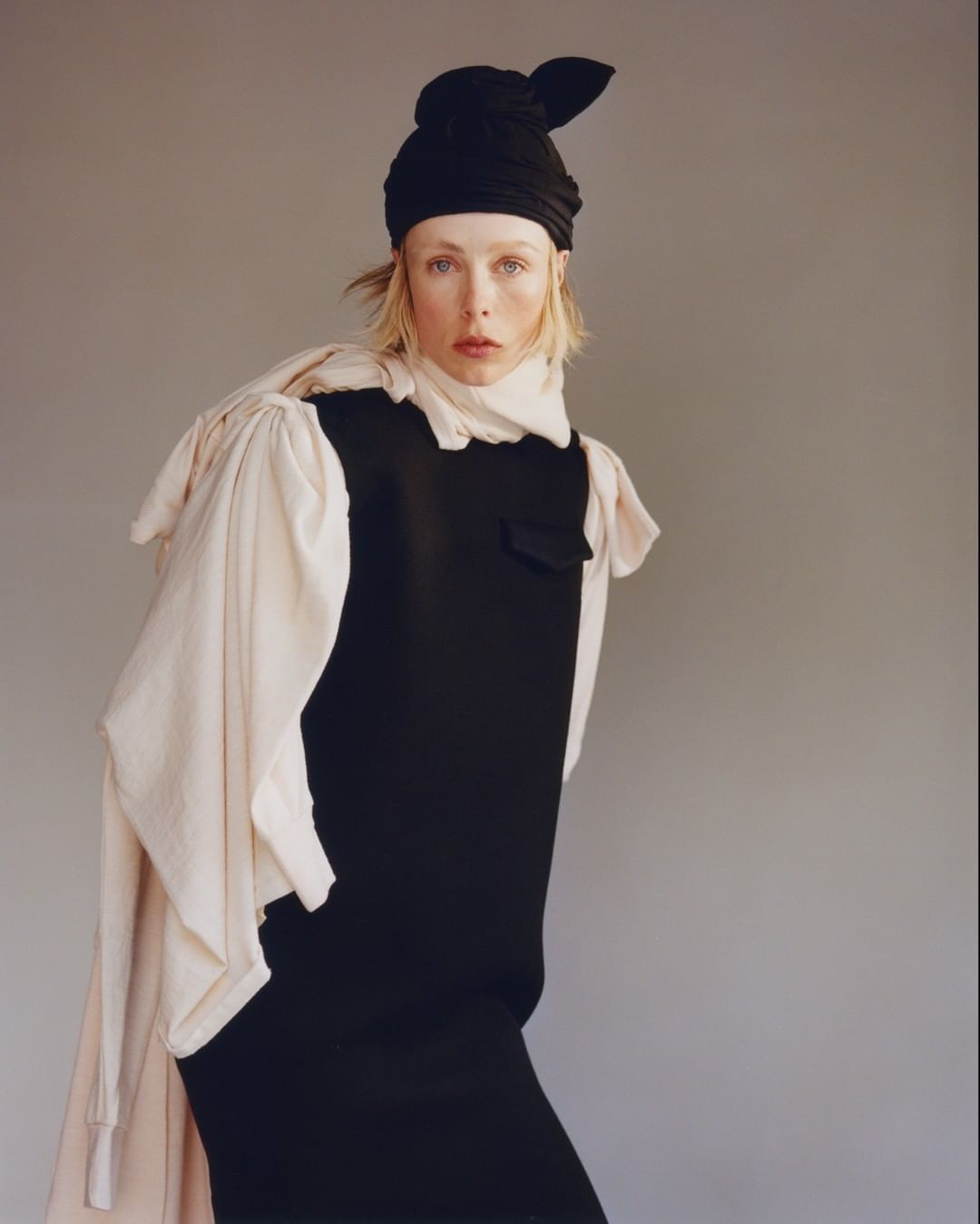 Fashion portrait of a stylish fair-skinned woman in a black dress and headwrap with cream-colored draped fabric, against a grey background.