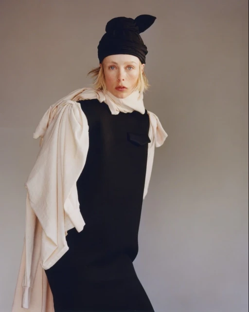 Fashion portrait of a stylish fair-skinned woman in a black dress and headwrap with cream-colored draped fabric, against a grey background.