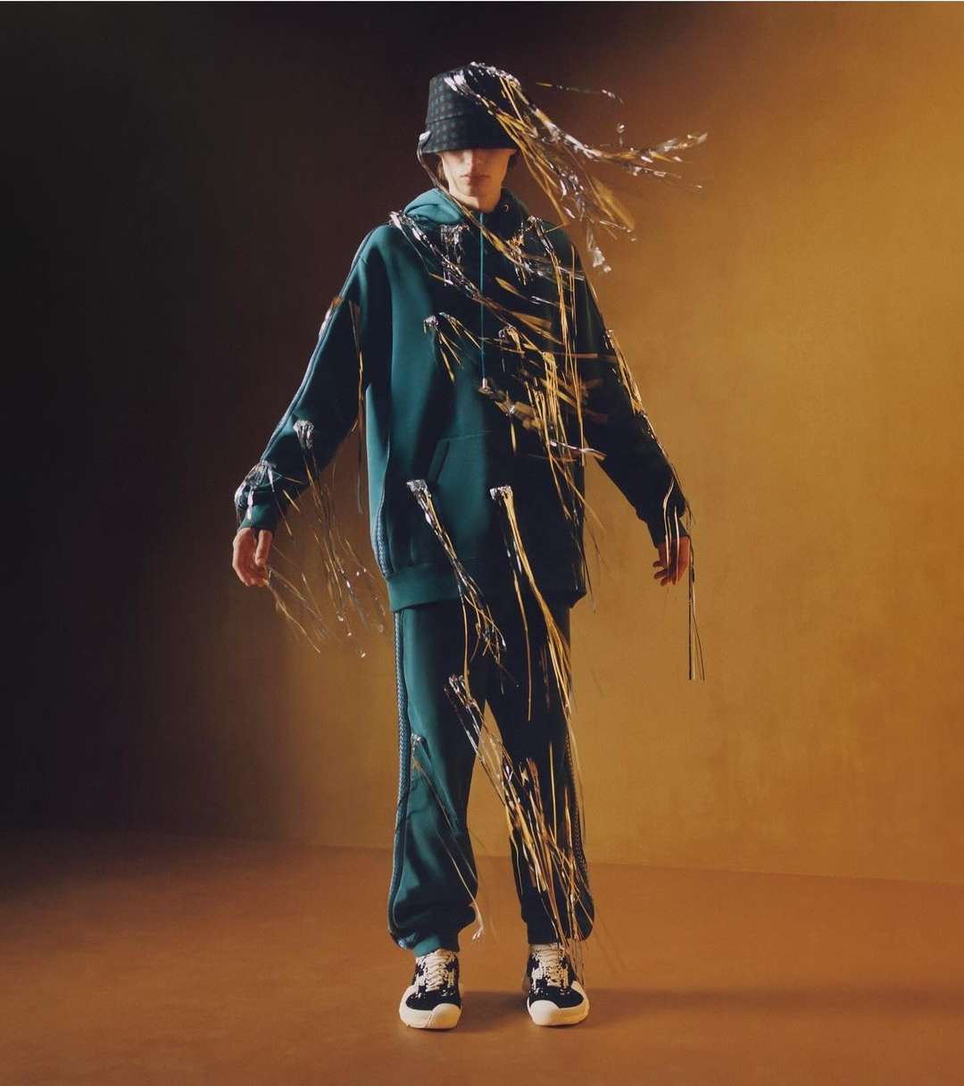 A full shot of a person wearing a matching green hooded sweatshirt and sweatpants with white and metallic gold decorations standing indoors.