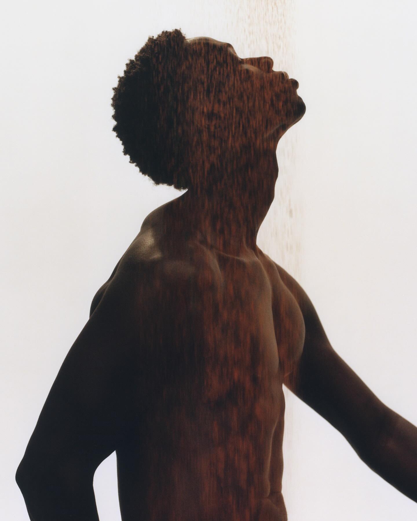 An artistic portrait of an African American male in a shower, looking upward with an expressive pose and dappled light on the body.