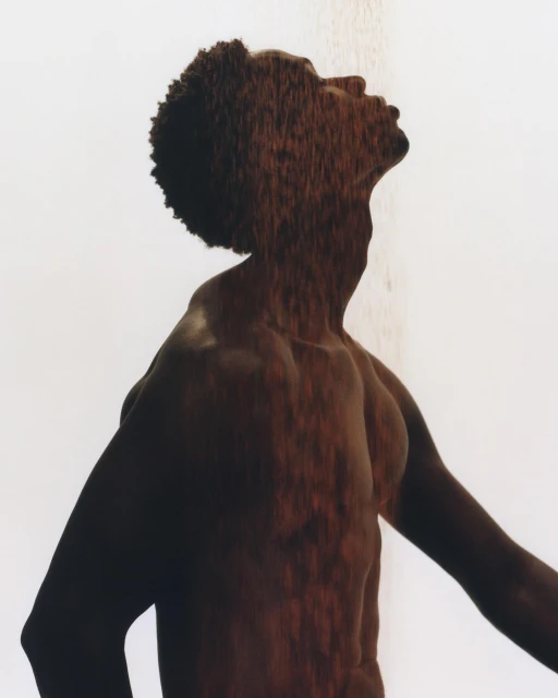 An artistic portrait of an African American male in a shower, looking upward with an expressive pose and dappled light on the body.
