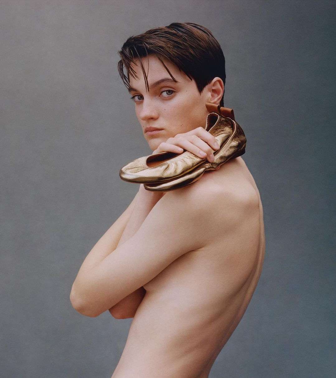 Portrait of a slender topless adult female holding a pair of gold ballet shoes over her shoulder against a solid grey backdrop.