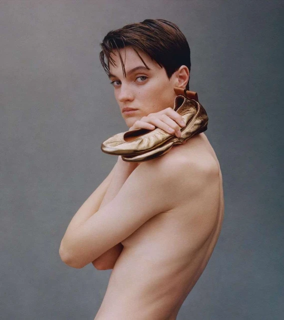 Portrait of a slender topless adult female holding a pair of gold ballet shoes over her shoulder against a solid grey backdrop.