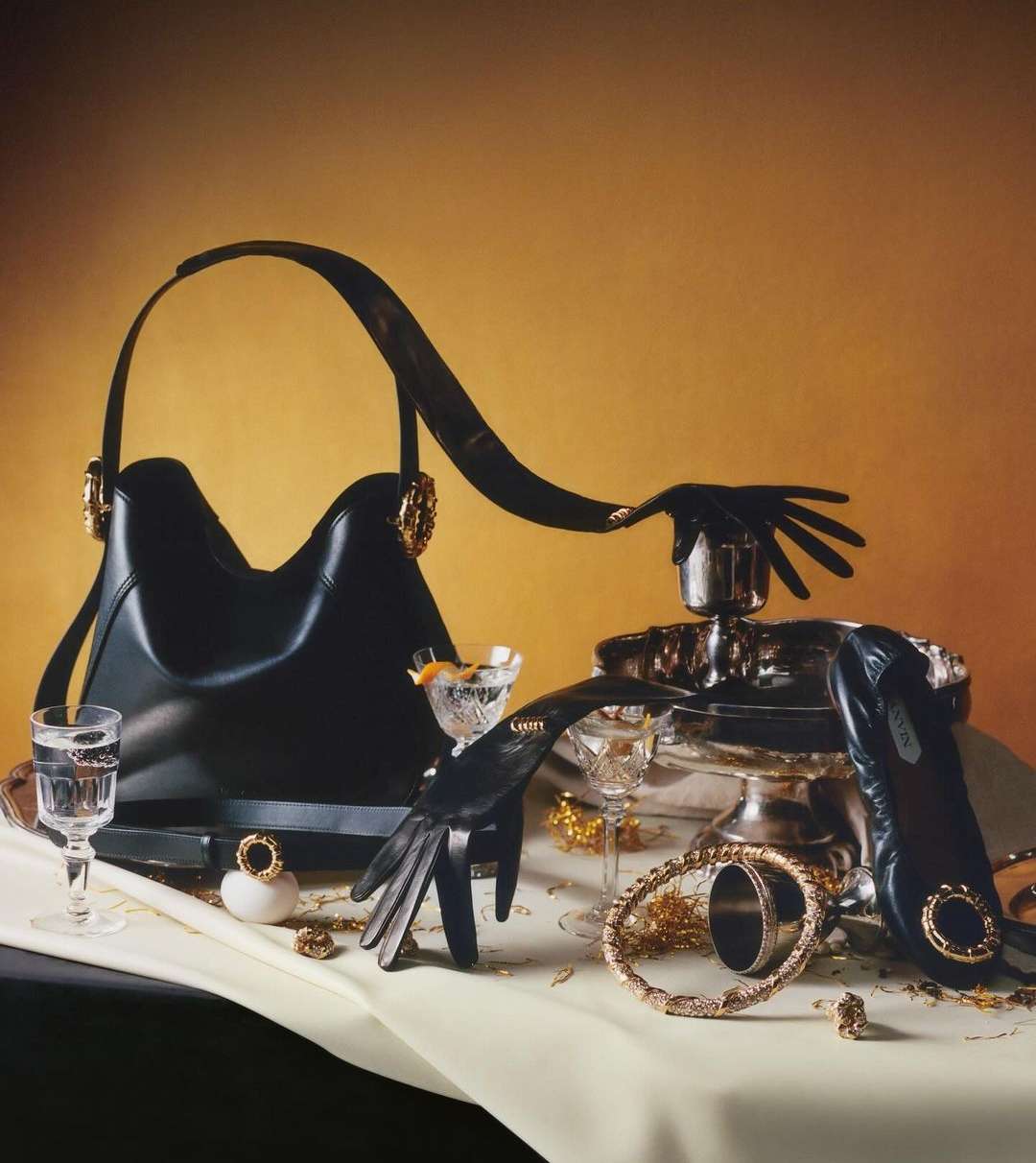 Still life shot of a black purse with gold hardware, cocktail glasses, gold jewelry, and black gloves on a white tablecloth.