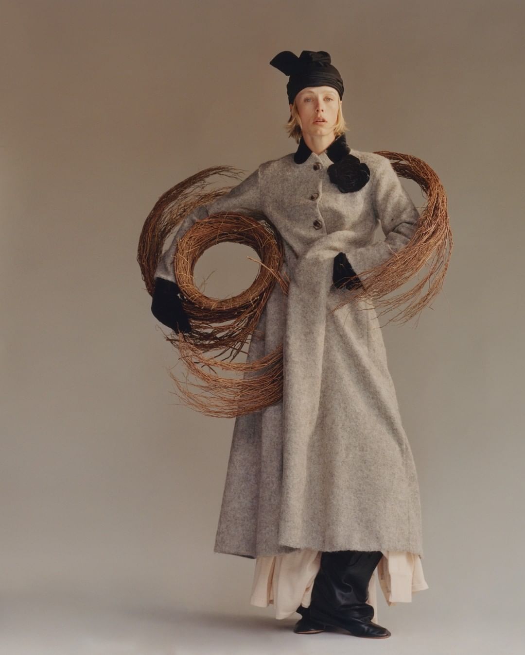 Model in grey tweed coat holding woven willow branches with black head wrap against a neutral studio background