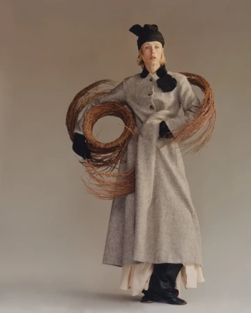 Model in grey tweed coat holding woven willow branches with black head wrap against a neutral studio background