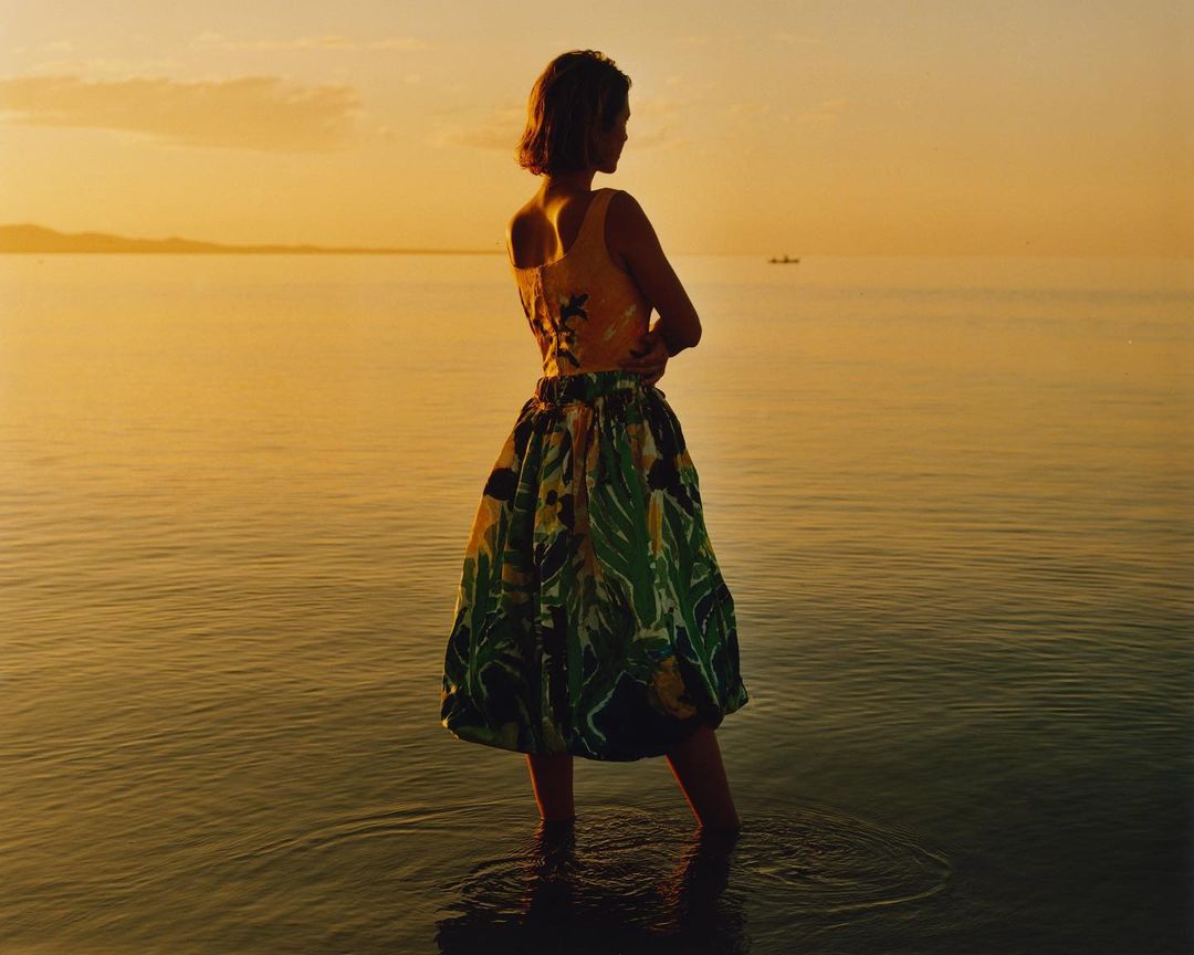 Woman in floral dress stands in the ocean at sunset, creating a serene and picturesque scene with warm lighting and a calm horizon.